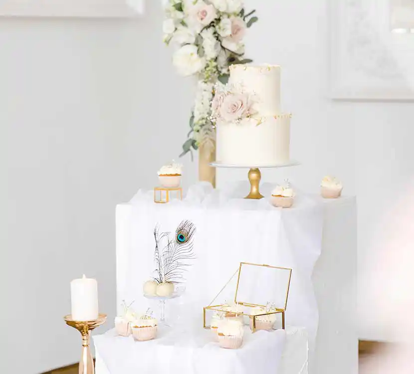 Weißer, gedeckter Tisch mit Hochzeitstorte, Cupcakes und Cakepops auf Teller mit Blumendeko.